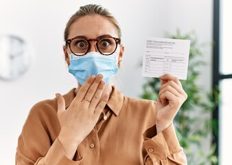 Young blonde woman holding covid record card covering mouth with hand, shocked and afraid for...