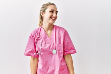 Young blonde woman wearing pink nurse uniform over isolated background looking away to side with...