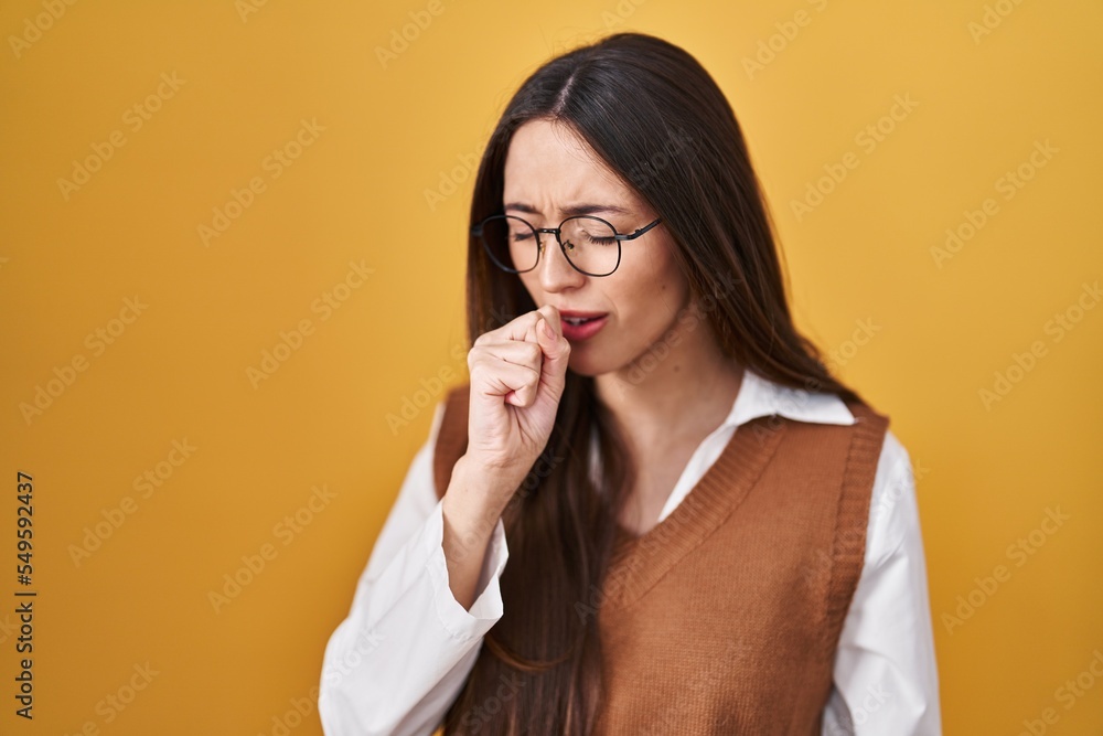 Sticker young brunette woman standing over yellow background wearing glasses feeling unwell and coughing as 