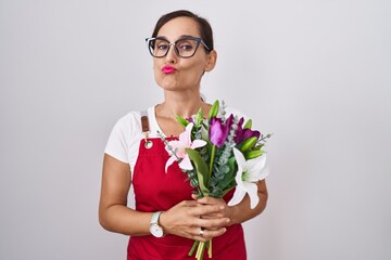 Middle age brunette woman wearing apron working at florist shop holding bouquet looking at the camera blowing a kiss on air being lovely and sexy. love expression.