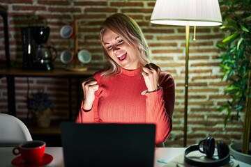 Blonde woman using laptop at night at home very happy and excited doing winner gesture with arms raised, smiling and screaming for success. celebration concept.