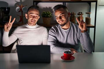 Homosexual couple using computer laptop smiling looking to the camera showing fingers doing victory...