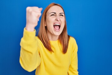 Young woman standing over blue background angry and mad raising fist frustrated and furious while shouting with anger. rage and aggressive concept.