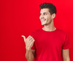 Young hispanic man standing over red background smiling with happy face looking and pointing to the side with thumb up.