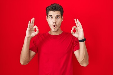 Fototapeta premium Young hispanic man standing over red background looking surprised and shocked doing ok approval symbol with fingers. crazy expression