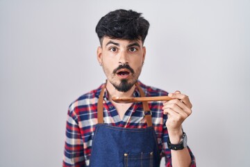 Young hispanic man with beard wearing apron tasting food holding wooden spoon scared and amazed...