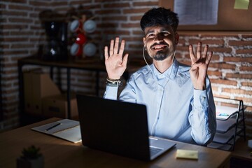 Young hispanic man with beard working at the office at night showing and pointing up with fingers number nine while smiling confident and happy.