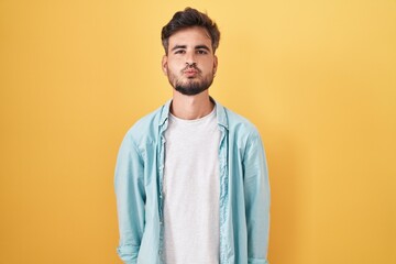 Young hispanic man with tattoos standing over yellow background looking at the camera blowing a...