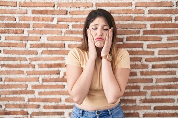 Young brunette woman standing over bricks wall tired hands covering face, depression and sadness, upset and irritated for problem