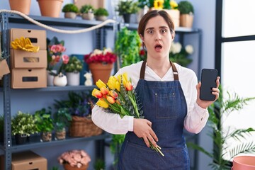 Brunette woman working at florist shop holding smartphone afraid and shocked with surprise and amazed expression, fear and excited face.