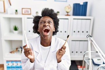 African dentist woman holding electric toothbrush and normal toothbrush angry and mad screaming...