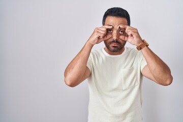 Hispanic man with beard standing over isolated background trying to open eyes with fingers, sleepy and tired for morning fatigue