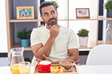 Hispanic man with beard eating breakfast thinking concentrated about doubt with finger on chin and looking up wondering