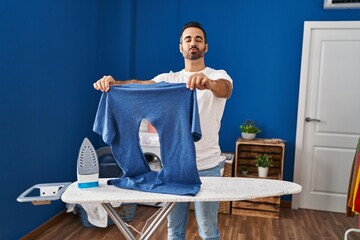 Young hispanic man with beard ironing holding burned iron shirt at laundry room looking at the...