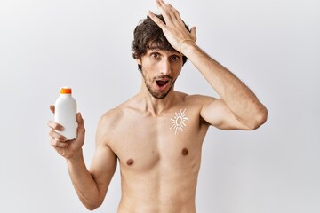 Fototapeta premium Young hispanic man standing shirtless holding sunscreen lotion surprised with hand on head for mistake, remember error. forgot, bad memory concept.