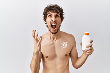 Fototapeta premium Young hispanic man standing shirtless holding sunscreen lotion crazy and mad shouting and yelling with aggressive expression and arms raised. frustration concept.