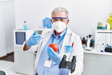 Middle age grey-haired man wearing scientist uniform holding syringe and apple at laboratory