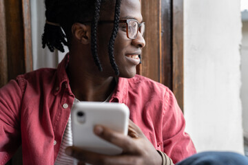 Positive satisfied African American man with smartphone smiling coming up with messages to become acquainted with girl. Joyful optimistic black guy with phone sits near window using social network