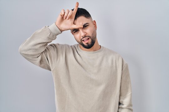 Young handsome man standing over isolated background making fun of people with fingers on forehead doing loser gesture mocking and insulting.