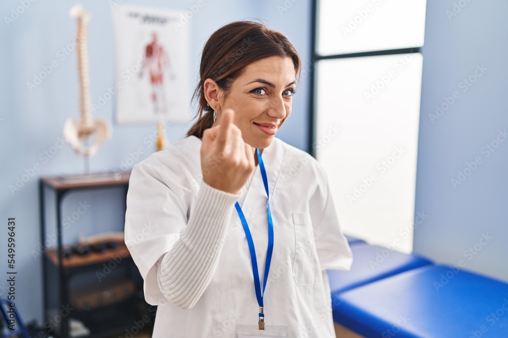 Poster young brunette woman working at pain recovery clinic beckoning come here gesture with hand inviting 