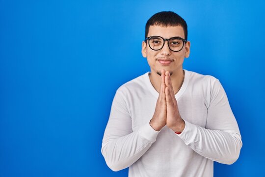 Young arab man wearing casual white shirt and glasses praying with hands together asking for forgiveness smiling confident.