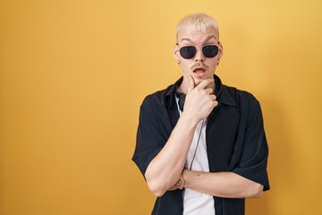 Young caucasian man wearing sunglasses standing over yellow background looking fascinated with disbelief, surprise and amazed expression with hands on chin