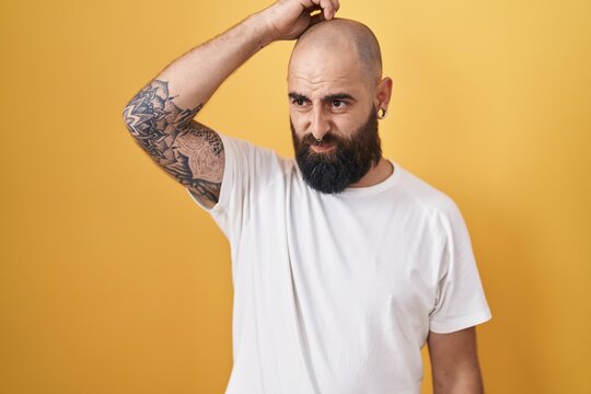 Young hispanic man with beard and tattoos standing over yellow background confuse and wondering about question. uncertain with doubt, thinking with hand on head. pensive concept.