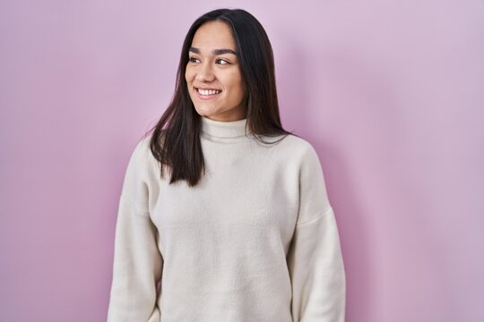 Young south asian woman standing over pink background looking away to side with smile on face, natural expression. laughing confident.