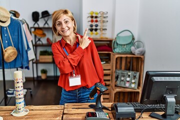 Middle age blonde woman working working as manager at retail boutique cheerful with a smile of face pointing with hand and finger up to the side with happy and natural expression on face