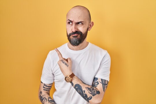 Young hispanic man with tattoos standing over yellow background pointing aside worried and nervous with forefinger, concerned and surprised expression