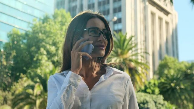 Hispanic businesswoman in glasses smiling speaks on a mobile outdoors. Funny brazilian young adult woman using a smart phone voice recognition. Pensive pretty young woman conversing on phone
