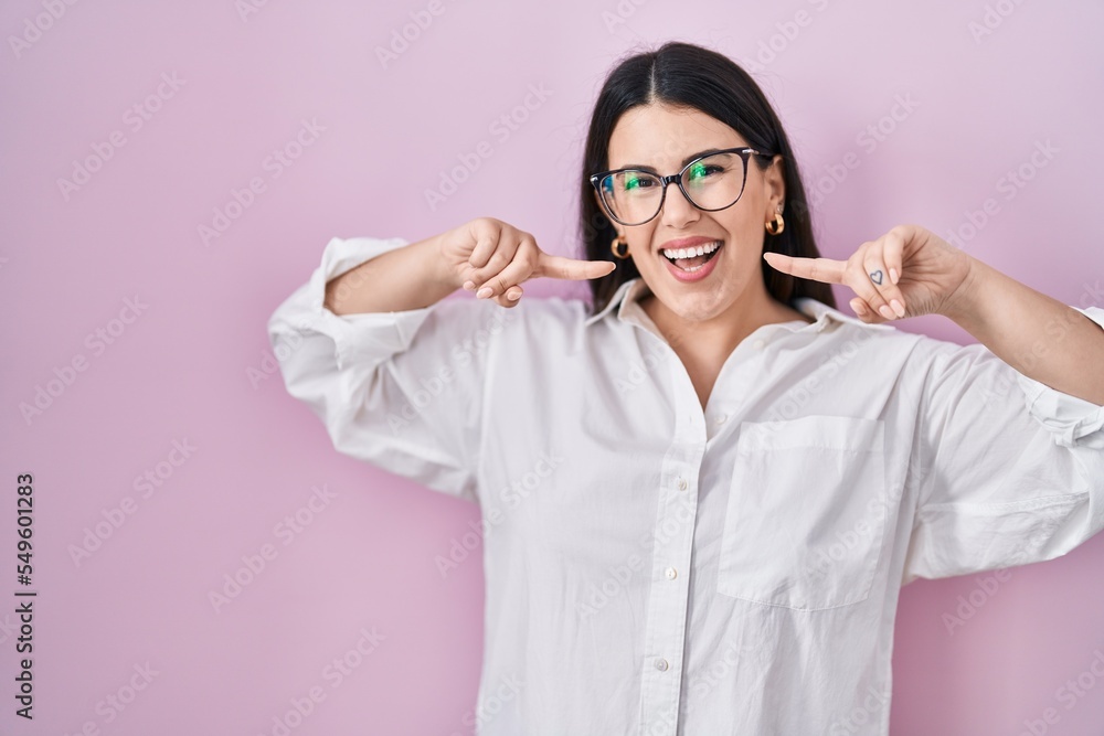 Sticker Young brunette woman standing over pink background smiling cheerful showing and pointing with fingers teeth and mouth. dental health concept.