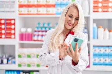 Young blonde woman pharmacist using smartphone working at pharmacy