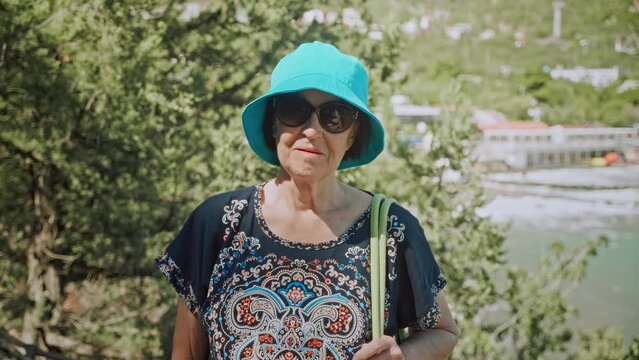 Happy and pretty elderly lady in sunglasses and blue panama hat, standing on hiking trail, posing for the camera. Summer vacation in warm country. Active lifestyle in retirement.