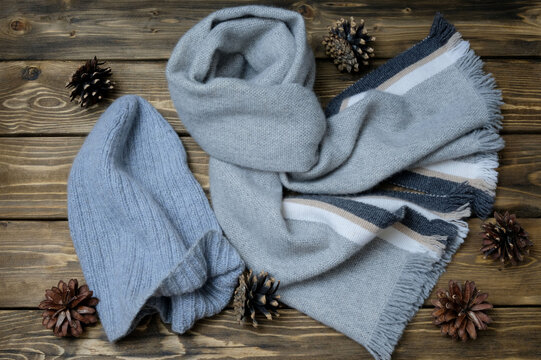 Striped grey cashmere scarf, tied in a knot.  The knitted cap is gray. Pine cones. All objects lie on the surface of dark-colored wooden boards.