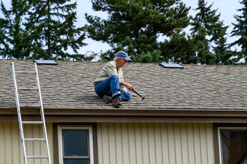 Senior man sitting on a house roof with a hammer, ready to make repairs
