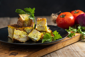 Pastel de papas y huevo cortado en cubos con perejil y tomate sobre plato negro 