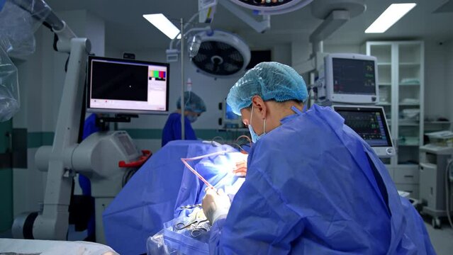 Back view of a doctor who bent over the patient conducting surgery. Neurosurgeon working in the modern surgical room with lots of advanced equipment.