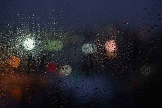 Raindrops on a window glass and unfocused lights of a night or evening city. Blurred Background of night city behind the glass during rain