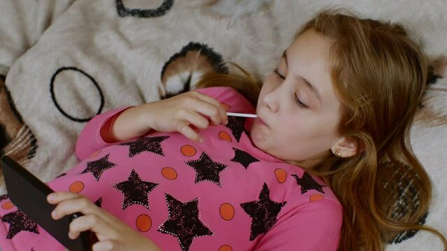 A girl with a smartphone in her left hand and a juicy lollipop in her right hand carefully looks at the smartphone screen and licks the lollipop. Concept of the problem of health and modern gadjit