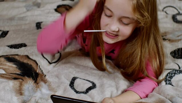 A cute little girl is lying on the couch, sucking on a sweet juicy lollipop, playing enthusiastically on her smartphone and raising her hand in victory. The concept of digital addiction in teenagers