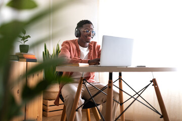 Long shot of young African American man wearing headphones working, studying at home using laptop....