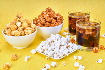 three bowls of spicy, caramelized and salted popcorn accompanied by 2 glasses of soda with ice on a yellow background with copy space for advertising.