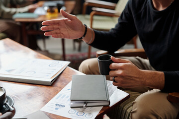Hands of young male economist with cup of tea sitting by table with notebook and financial documents during conversation with colleague