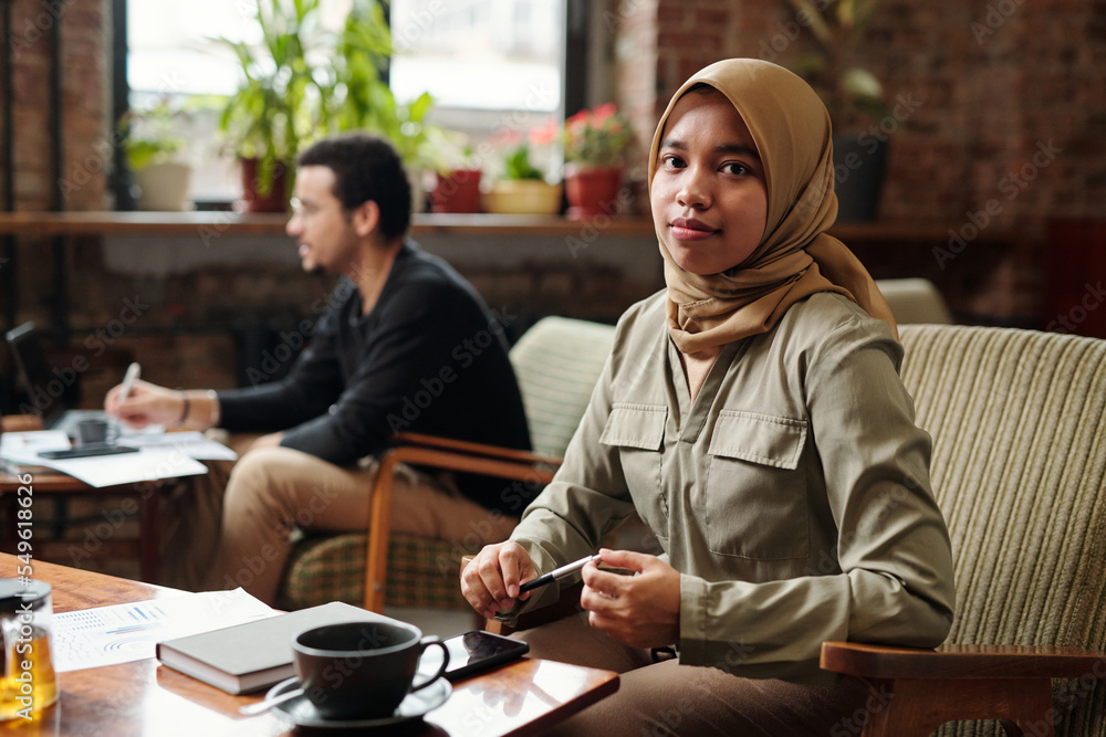 Poster young pretty businesswoman in hijab sitting by table in front of camera and looking at you while org