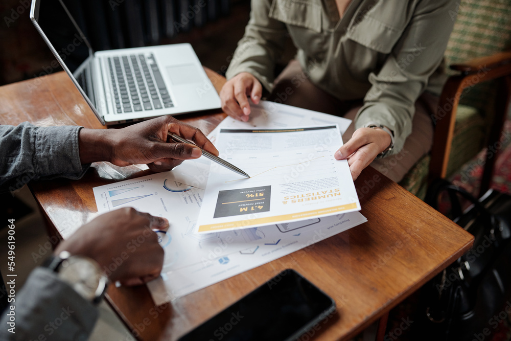 Poster hand of young black man with pen pointing at financial document while explaining data to female coll