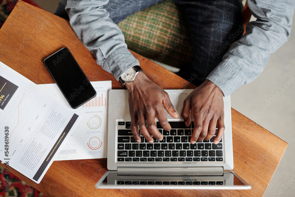 Poster top view of hands of young black man pushing keys of laptop keyboard while sitting by desk with smar