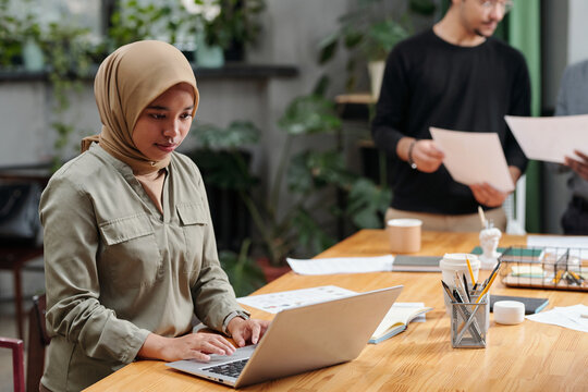 Young serious businesswoman in hijab looking at laptop screen while sitting by workplace and organizing work against colleagues