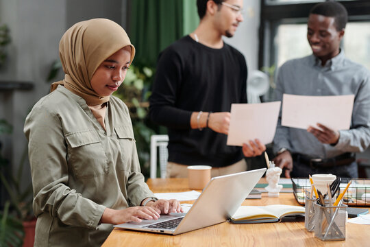 Young Muslim businesswoman organizing work in front of laptop while standing by workplace against two interculural men with papers