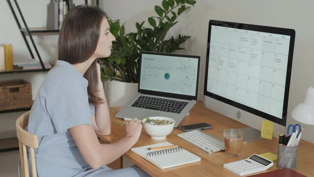 Young accountant having homemade food from bowl while sitting in front of computer screen and looking through spreadsheet with data